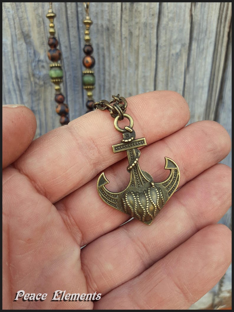 May include: A bronze-toned metal anchor pendant featuring a textured rope design on the fluke, suspended from a chain. The necklace includes a strand of brown and green beads. The text Peace Elements is visible in the bottom left corner.