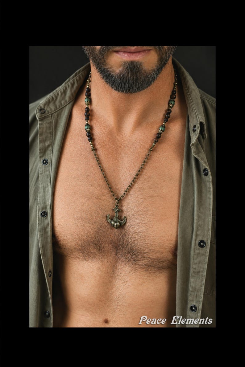 May include: A necklace featuring a bronze-toned anchor pendant suspended from a chain accented with dark brown wooden beads and small green beads. The text Peace Elements is visible in the bottom right corner of the image.
