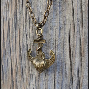 May include: A bronze-toned metal necklace featuring a pendant shaped like a nautical anchor combined with a heart design. The pendant hangs from a matching thick oval-link chain. The text Peace Elements is visible in the bottom right corner.