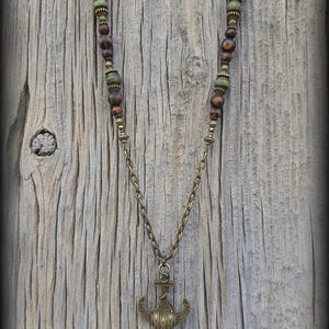 May include: A necklace featuring a bronze-toned anchor pendant suspended from a chain. The upper portion of the necklace consists of brown mottled beads and smaller green beads separated by metallic spacers. The text Peace Elements is visible.