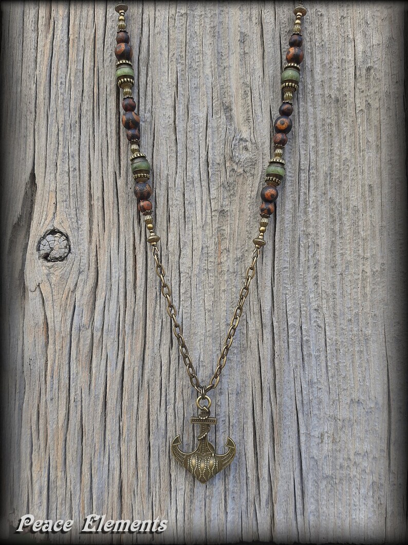 May include: A necklace featuring a bronze-toned anchor pendant suspended from a chain. The upper portion of the necklace consists of brown mottled beads and smaller green beads separated by metallic spacers. The text Peace Elements is visible.