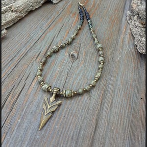 Op de afbeelding: Een handgemaakte ketting met een bronzen pijlhanger en een kralenketting. De kralen zijn een mix van groen, grijs en brons, met een donkerblauwe accent op de sluiting. De ketting ligt op een verweerd houten oppervlak. De tekst "Peace Elements" is zichtbaar.