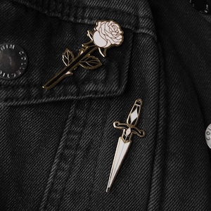 May include: A black denim jacket with two gold pins. One pin is a white rose with a gold stem and leaves. The other pin is a gold dagger with a white blade.