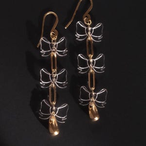 May include: Gold-tone drop earrings featuring a series of three clear bow-shaped charms linked by gold-tone bars. The earrings have a gold-tone hook and a teardrop-shaped charm at the bottom. The earrings are displayed against a black background.