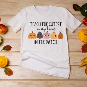May include: White t-shirt with a cute graphic of pumpkins with faces and the text "I teach the cutest pumpkins in the patch". The shirt is surrounded by fall-themed decorations, including gourds, leaves, and pumpkins.