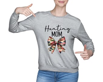 Op de afbeelding: Een grijze longsleeve sweatshirt met de tekst "Hunting MOM" in zwart script. Een camouflage strik grafisch is gecentreerd onder de tekst. De sweatshirt wordt gecombineerd met een blauwe jeans.