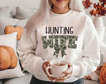 Op de afbeelding: Wit sweatshirt met de tekst "HUNTING WIFE" in zwarte letters met een camouflagepatroon. Het woord "WIFE" heeft eronder een camouflage strik.