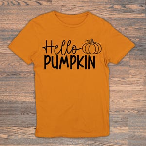 May include: An orange t-shirt featuring the text "Hello Pumpkin" in black script and block letters, accompanied by a pumpkin graphic. The shirt is shown on a wooden background.