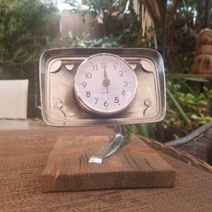 May include: A silver clock with a white face and black numbers is mounted on a silver car part. The car part is attached to a wooden base.