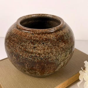 May include: A round, squat ceramic vase with a wide opening and a thick, rolled rim. The exterior features a mottled, textured glaze in earthy tones of brown, tan, and dark gray, with visible speckling and horizontal striations across the surface.