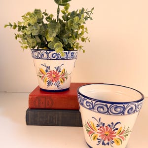 May include: Two white ceramic flower pots featuring hand-painted blue rim detailing with swirling patterns and a central floral motif in pink, yellow, and green. One pot contains a green leafy plant, while the other sits empty in front.