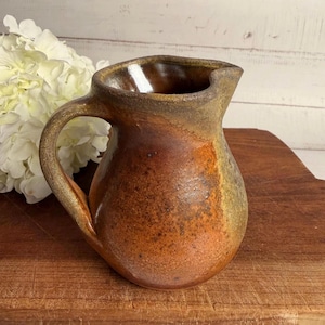 May include: A handmade ceramic pitcher with a rustic, earthy aesthetic. The pitcher features a warm color palette of browns and oranges, with a rounded body, a curved handle, and a pouring spout. The pitcher is displayed on a wooden surface.