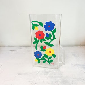 May include: A clear glass pitcher with a floral design. The flowers are red, blue, yellow, and green. The pitcher has a handle and a label that says "GS".