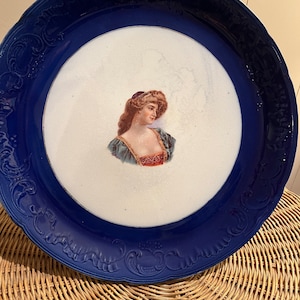 May include: A blue and white porcelain plate with a raised rim and a decorative border. The center of the plate features a painted image of a woman with a crown.
