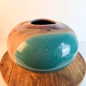 May include: A round ceramic vase with a wide, low-profile body and a small circular opening. The surface features a glossy glaze with a distinct crackle pattern, transitioning from a deep teal green on the lower half to a muted tan and purple.