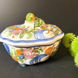 May include: A ceramic trinket box with a lid, featuring a floral design in pink, orange, blue, and green. The box has a blue border and a decorative knob on top. The overall shape is rounded and ornate.