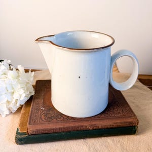 May include: A white ceramic creamer pitcher with a speckled finish and a brown rim. The pitcher features a curved handle on the right side and a pouring spout on the left. The surface of the pitcher has small dark speckles throughout the body.