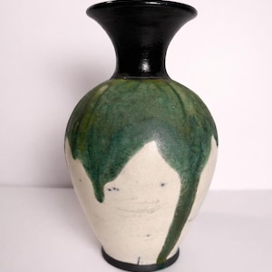 May include: A ceramic vase with a black rim and base, and a white body. The vase is decorated with green glaze that drips down the sides. The vase has a rounded body and a flared neck.