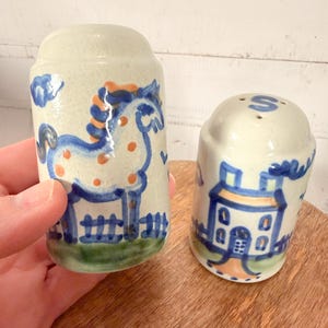 May include: A pair of ceramic salt and pepper shakers. One shaker features a blue horse design with orange accents and a green base. The other shaker has a house design with the letter "S" on top.