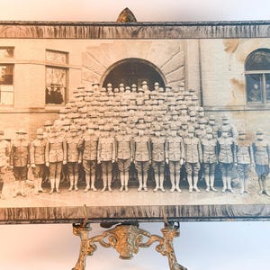 May include: A rectangular framed black and white photograph depicting a large group of soldiers in uniform standing in rows in front of a stone building. The frame has a dark, wood-grain pattern and is supported by an ornate gold metal stand.
