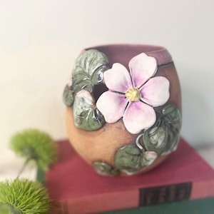May include: A ceramic vase with a light brown exterior and a dark purple interior. The vase is adorned with a large, three-dimensional pink flower and green leaves. The vase is sitting on top of a red book.