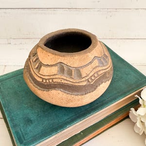 May include: A round, tan-colored ceramic bowl with a wide opening and a dark, textured interior. The exterior features a rough, sandy surface with horizontal bands of dark brown, carved geometric patterns and vertical indentations.