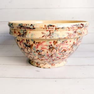 May include: A large, cream-colored ceramic mixing bowl with a speckled pattern of red, blue, and brown. The bowl has a wide rim and a pedestal base, suitable for kitchen use.