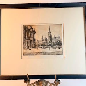 May include: A black-framed etching featuring a detailed architectural scene of a large gothic-style building with a tall central spire, surrounded by trees and figures in a plaza, set within a cream-colored mat board with a signature in the corner.