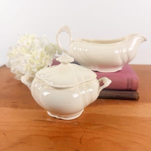 May include: A cream-colored ceramic sugar bowl with a matching lid and a cream-colored gravy boat. Both pieces feature a smooth, glossy finish, subtle vertical fluting, and delicate, ornate handles with small floral-like detailing.