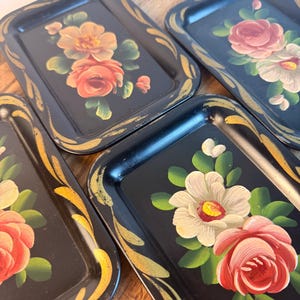May include: A set of three rectangular black metal trays featuring hand-painted floral motifs in shades of pink, white, and green. Each tray is decorated with gold-toned brushstroke accents along the edges and displays a unique arrangement of roses.