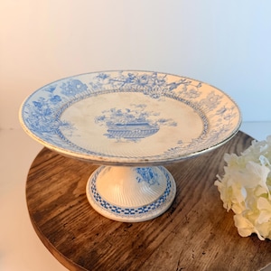 May include: A ceramic pedestal cake stand featuring a white base with blue floral and botanical patterns. The circular plate has a scalloped edge and a central urn motif, while the flared base is decorated with a blue and white checkered border.