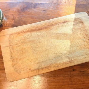 May include: A rectangular light-colored wooden cutting board with rounded corners and a smooth surface featuring visible knife marks and scratches from use. The board has a natural grain pattern and is positioned on a polished wooden surface.