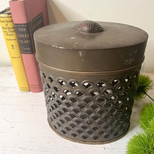 May include: A vintage brown metal container with a lid and a decorative shell-shaped handle. The container has a patterned, openwork design. Three antique books are visible in the background.