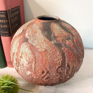 May include: A round, textured ceramic vase with a dark brown interior and a mottled exterior in shades of red, brown, and cream. The vase has a wide opening and sits on a white surface.