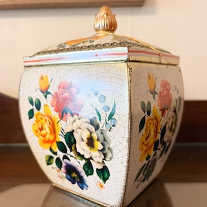 May include: A white square tin box with a gold lid and a floral design. The lid has a gold knob on top. The box is decorated with a floral pattern of yellow, pink, white, and blue flowers.