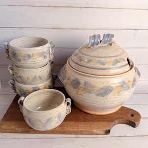 May include: A stoneware soup tureen with lid and matching bowls. The set features a speckled beige glaze with blue and tan leaf-like designs. The tureen has handles and a ladle opening. The bowls have double handles.