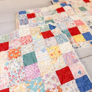 May include: A set of two square quilted potholders featuring a patchwork design of small fabric squares in various colors including red, blue, yellow, green, and floral patterns. The quilted fabric has visible stitching and a white fabric border.