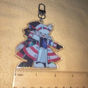Puede incluir: Un llavero de acr&iacute;lico transparente con un personaje de dibujos animados con abrigo blanco, sombrero y detalles en rojo, blanco y azul. El llavero mide aproximadamente 5 cm de alto y tiene un cierre de metal.