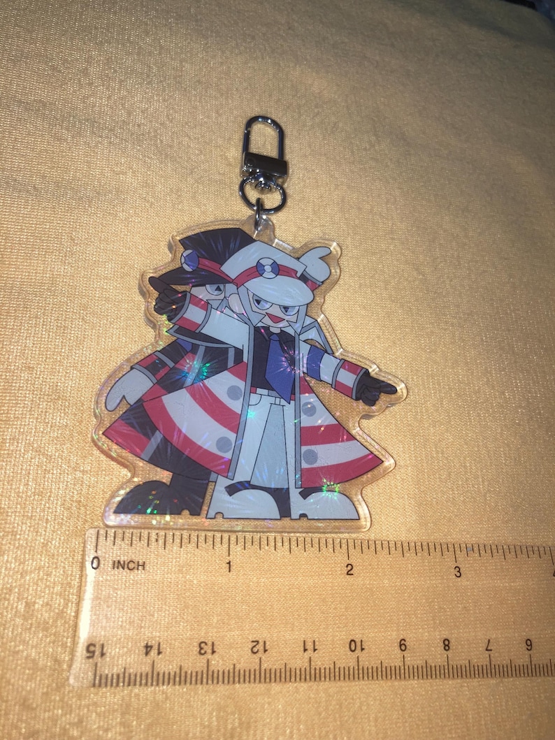 Puede incluir: Un llavero de acr&iacute;lico transparente con un personaje de dibujos animados con abrigo blanco, sombrero y detalles en rojo, blanco y azul. El llavero mide aproximadamente 5 cm de alto y tiene un cierre de metal.