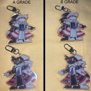 Puede incluir: Cuatro llaveros con un personaje de dibujos animados con un atuendo negro, blanco y rojo. Cada llavero tiene un cierre plateado. Los textos "A GRADE" y "B GRADE" son visibles encima de los llaveros.