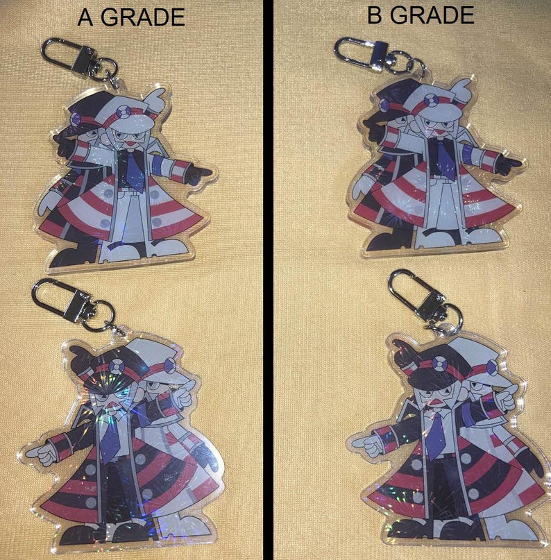 Puede incluir: Cuatro llaveros con un personaje de dibujos animados con un atuendo negro, blanco y rojo. Cada llavero tiene un cierre plateado. Los textos "A GRADE" y "B GRADE" son visibles encima de los llaveros.
