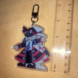 Puede incluir: Un llavero acr&iacute;lico transparente con una ilustraci&oacute;n de dibujos animados de dos figuras con trajes a juego. Las figuras llevan sombreros, abrigos y corbatas. El llavero est&aacute; sujeto a un cierre plateado. La regla mide 10 cm.