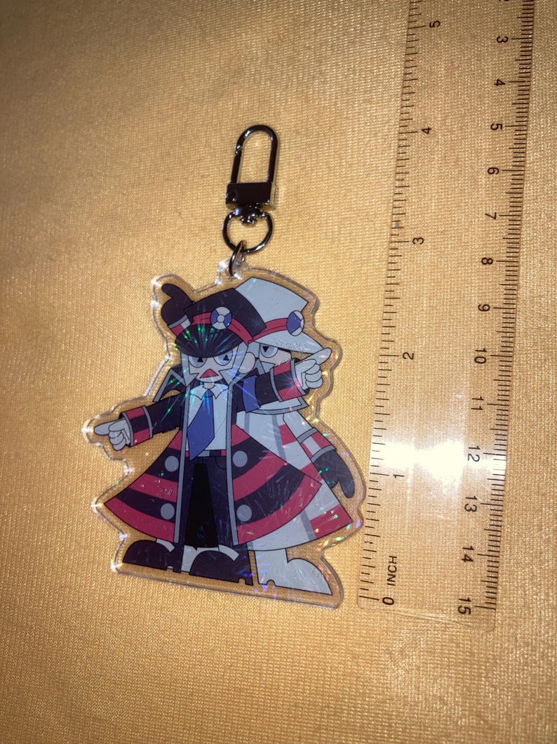 Puede incluir: Un llavero acr&iacute;lico transparente con una ilustraci&oacute;n de dibujos animados de dos figuras con trajes a juego. Las figuras llevan sombreros, abrigos y corbatas. El llavero est&aacute; sujeto a un cierre plateado. La regla mide 10 cm.