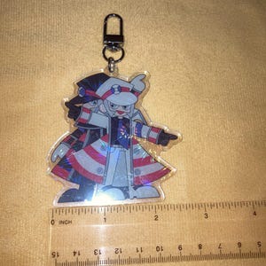 Puede incluir: Llavero acr&iacute;lico transparente con un personaje de dibujos animados con un abrigo con detalles en rojo, blanco y azul. El llavero tiene un cierre plateado. El personaje mide unos 5 cm de largo.