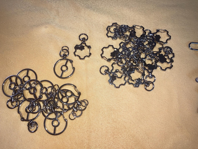 Puede incluir: Varios dijes y conectores met&aacute;licos plateados est&aacute;n dispuestos sobre una superficie beige. Los dijes presentan dise&ntilde;os circulares y en forma de engranaje, conectados por peque&ntilde;os anillos. Son componentes para joyer&iacute;a o manualidades.