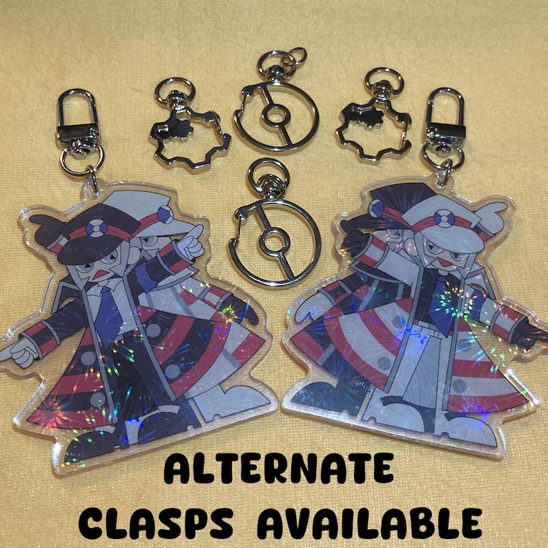 Puede incluir: Dos llaveros acr&iacute;licos con un personaje de dibujos animados con un atuendo negro, rojo y blanco, con sombrero y corbata a juego. Los llaveros tienen cierres plateados. El texto "ALTERNATE CLASPS AVAILABLE" est&aacute; en la parte inferior.