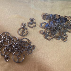 Puede incluir: Una colecci&oacute;n de dijes y colgantes de metal plateado, que incluyen dise&ntilde;os de c&iacute;rculos abiertos y piezas en forma de flor. Los dijes est&aacute;n dispersos sobre una superficie de color amarillo claro, ofreciendo una variedad de formas y tama&ntilde;os.