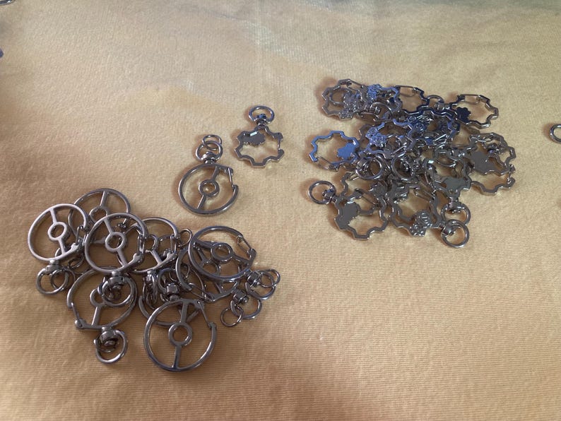 Puede incluir: Una colecci&oacute;n de dijes y colgantes de metal plateado, que incluyen dise&ntilde;os de c&iacute;rculos abiertos y piezas en forma de flor. Los dijes est&aacute;n dispersos sobre una superficie de color amarillo claro, ofreciendo una variedad de formas y tama&ntilde;os.