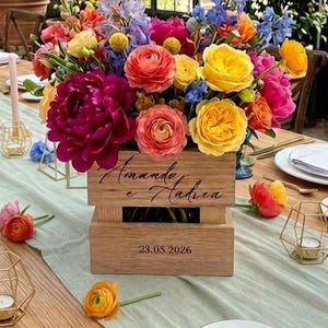 Op de afbeelding: Een houten bloemstuk met levendige bloemen in roze, geel, oranje en blauw. De kist heeft tekst en de datum 23.05.2026. De tafel is gedekt met glaswerk, borden en gouden kandelaars, wat een elegante sfeer creëert.