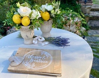 May include: A wooden guest book with the inscription "Alessio & Beatrice 01.08.2020" in white lettering. The book is decorated with a heart-shaped wooden tag and is placed on a white tablecloth. Two white ceramic vases with lemon blossoms and lemons are in the background.